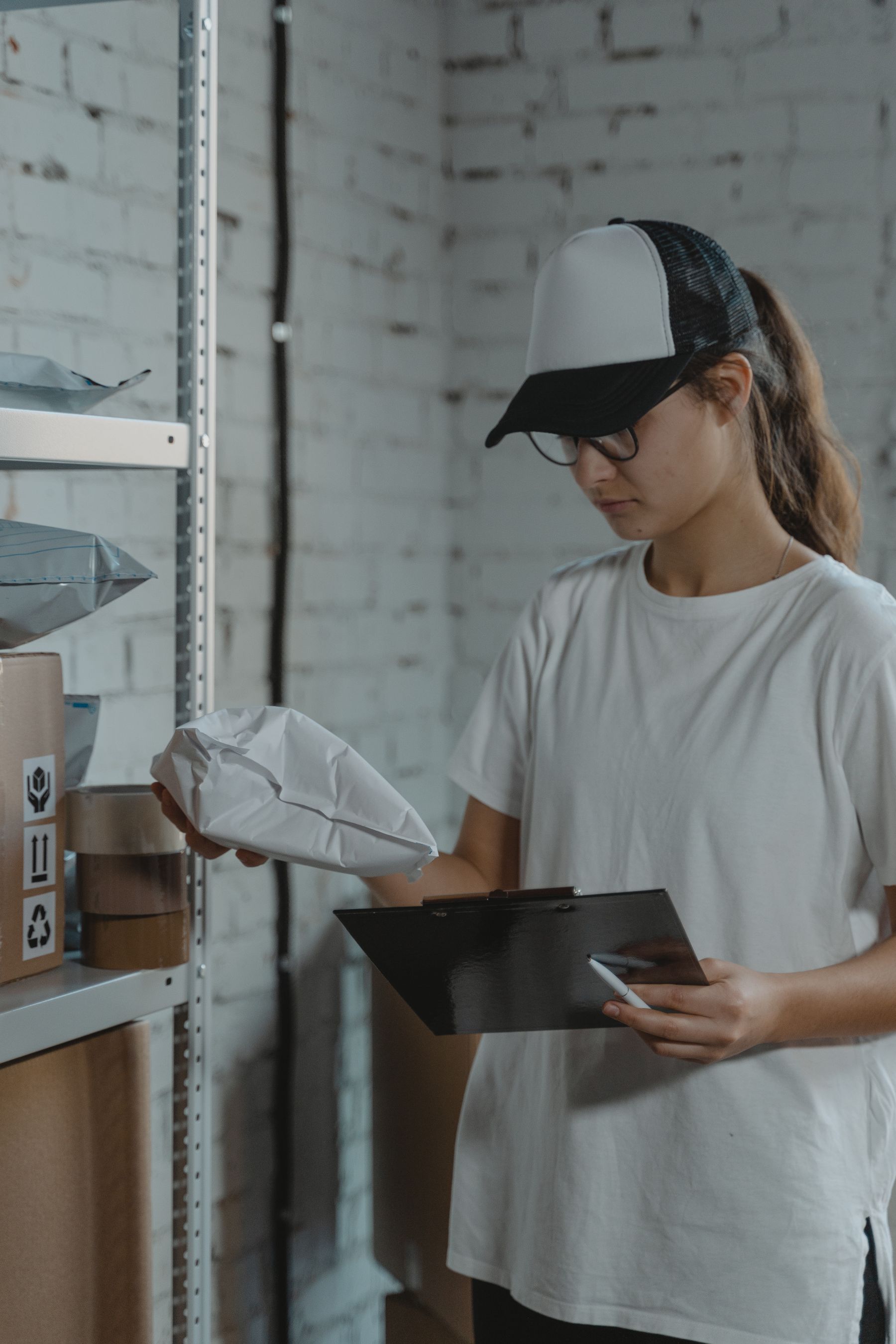 Warehouse worker in a white shirt and cap checks a package while holding a clipboard