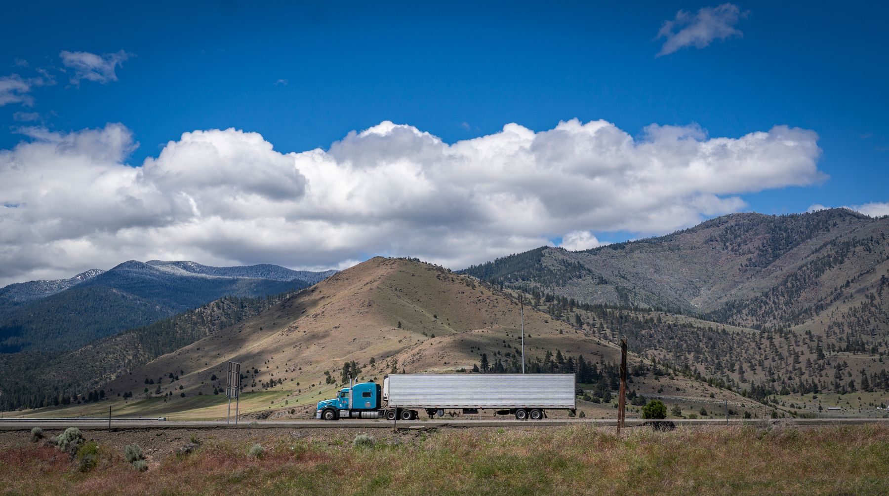 Blue semi-truck hauling a box trailer past mountain foothills on a western highway